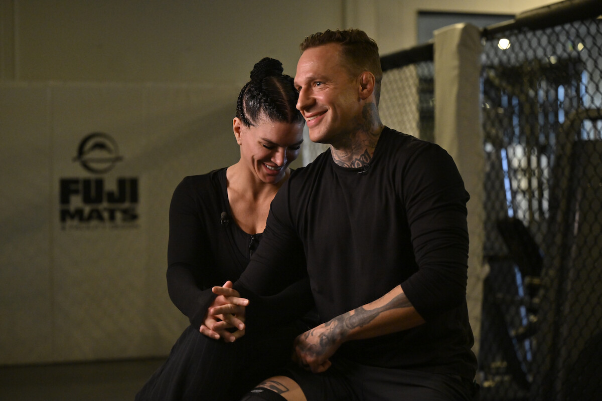 Two people sitting and smiling together inside an MMA gym, with a FUJI MATS logo on the wall and part of an octagon cage visible in the background, both dressed in black athletic wear.