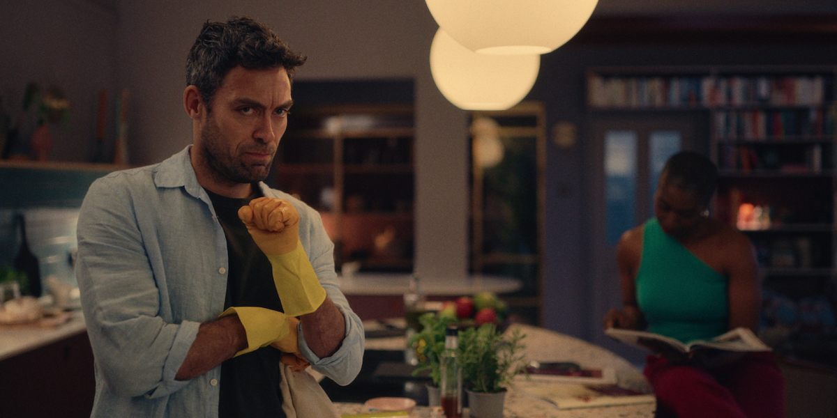 Alex Hassell as Rick stands in a kitchen wearing yellow rubber gloves in Season 1 of ‘Everything Now.’