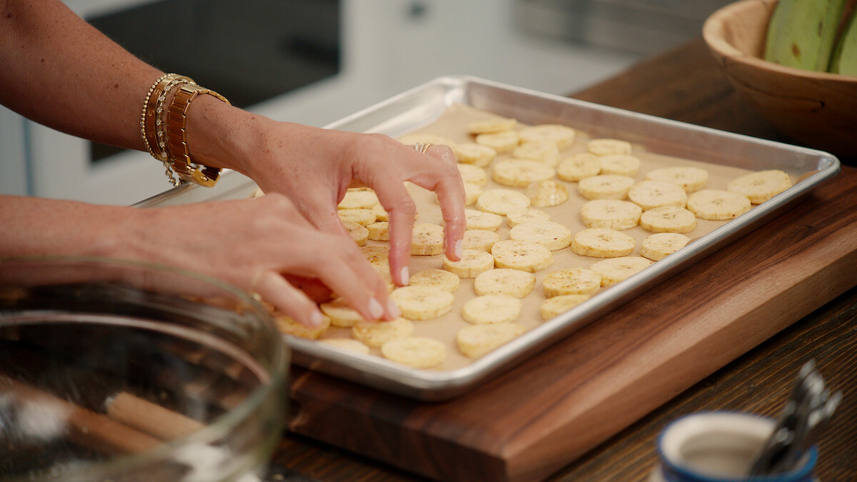 Sliced plantains being placed on a baking sheet.