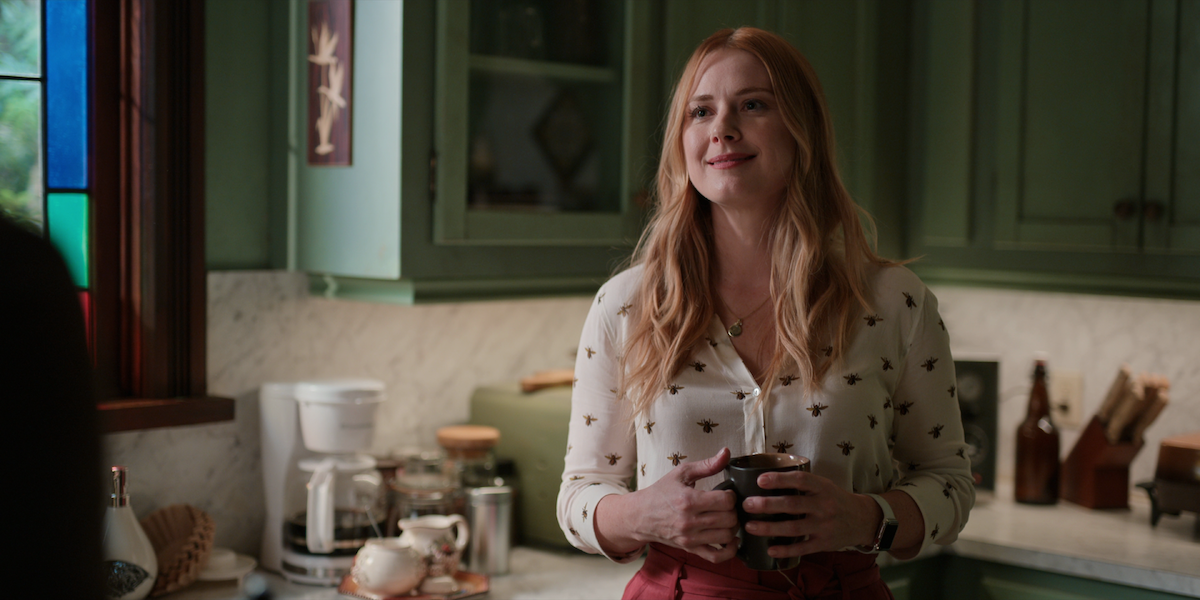 Alexandra Breckenridge holding a mug of coffee smirking at someone off screen.