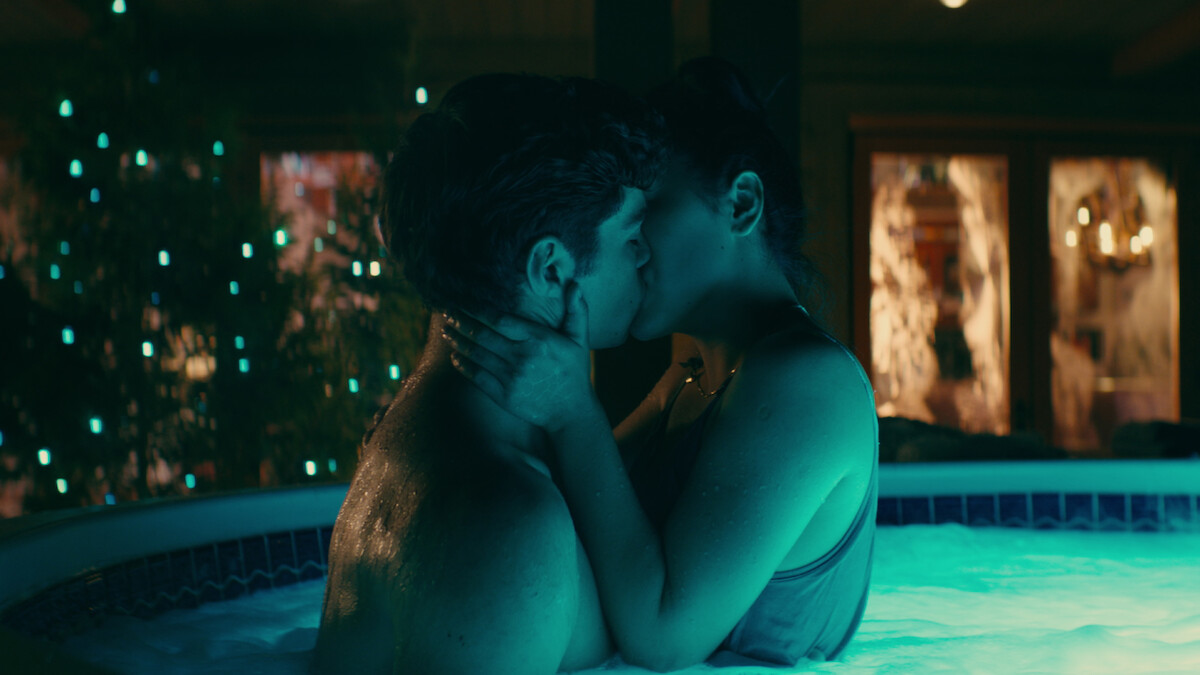 A couple kissing in a hot tub at night, illuminated by blue and green pool lights, with a decorated tree and house windows in the background.