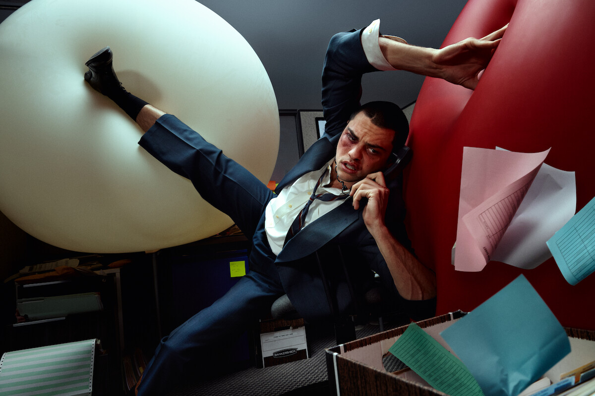  A beaten up looking Noah Centineo on a cell phone, in an office while his legs are resting on large inflatable balloons. 