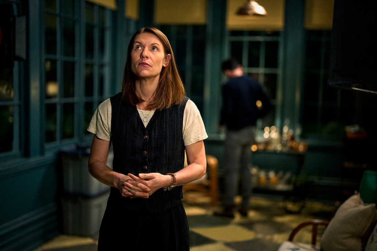 A woman stands indoors in a warmly lit room with large windows, looking thoughtful. In the background, a man faces away, suggesting a quiet, contemplative or tense mood. The setting appears cozy and intimate.