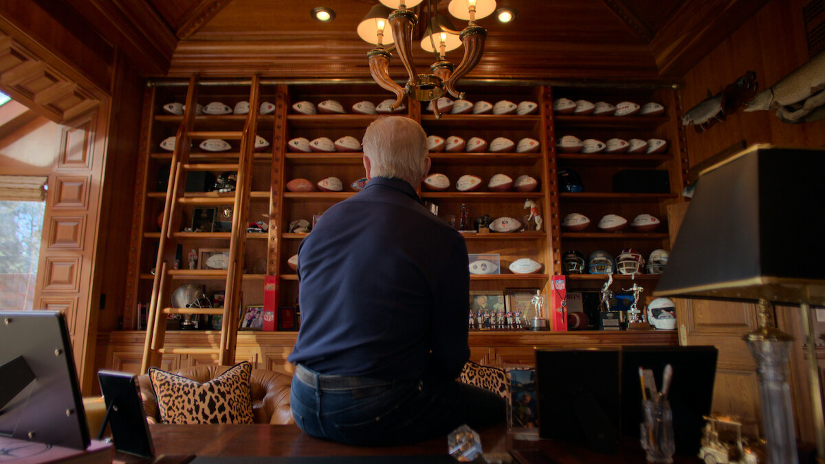 John Elway sits on a desk in a wood-paneled room facing shelves filled with football helmets, signed footballs, and trophies, with a ladder and animal mounts also visible in a well-lit study or office environment.