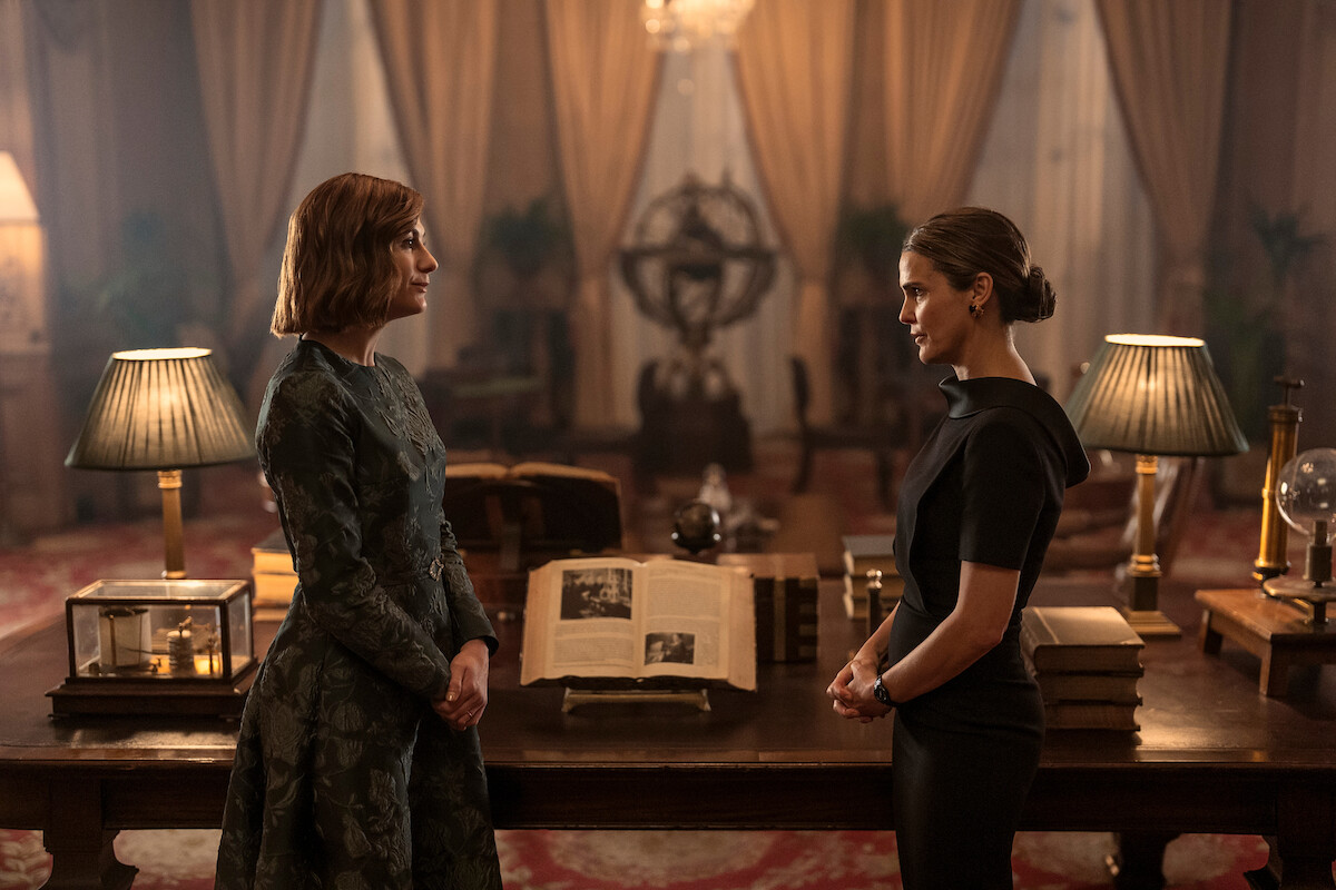 Two women stand facing each other in an elegant, dimly lit library or study, with books, lamps, and large curtains in the background, creating a serious and formal atmosphere.