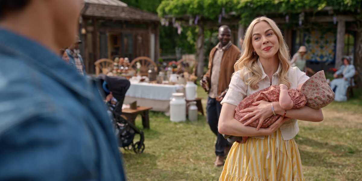 A woman in a yellow skirt holds a baby at an outdoor gathering, smiling and talking to a man in the foreground, with tables, people, and rustic decorations in the background on a sunny day.
