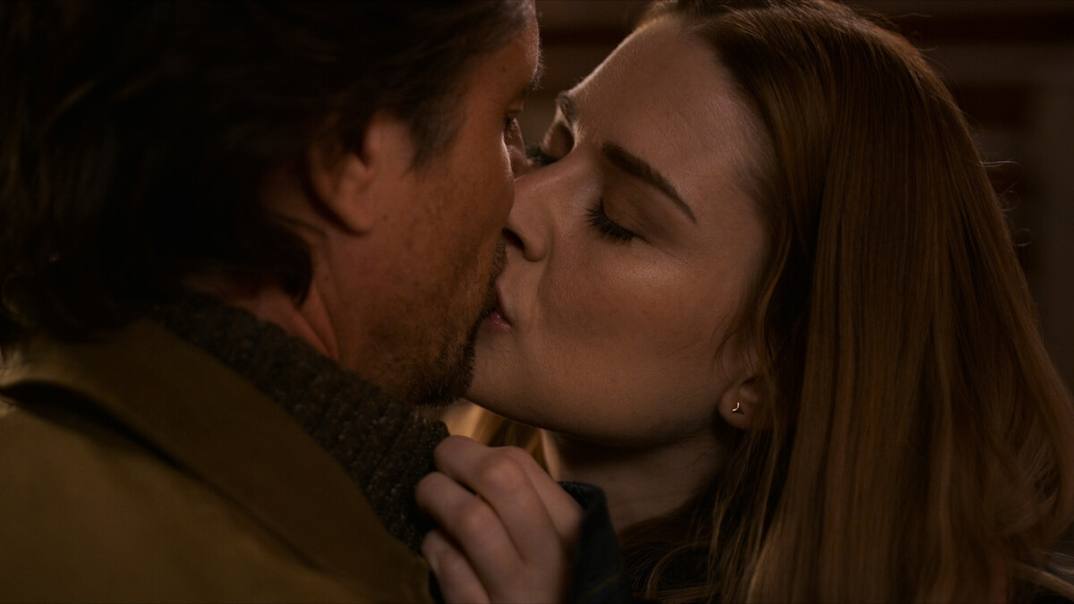 A man and a woman share a passionate kiss indoors, suggesting a moment of intimacy and affection. Warm lighting and close framing enhance the personal and emotional atmosphere.