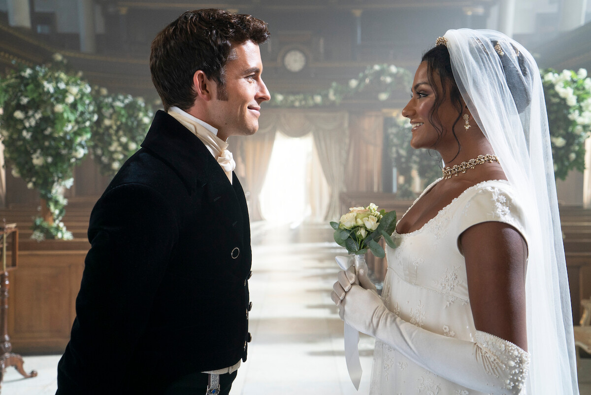 A bride and groom in formal attire smiling at each other inside a flower-decorated, sunlit wedding venue with elegant wooden architecture.