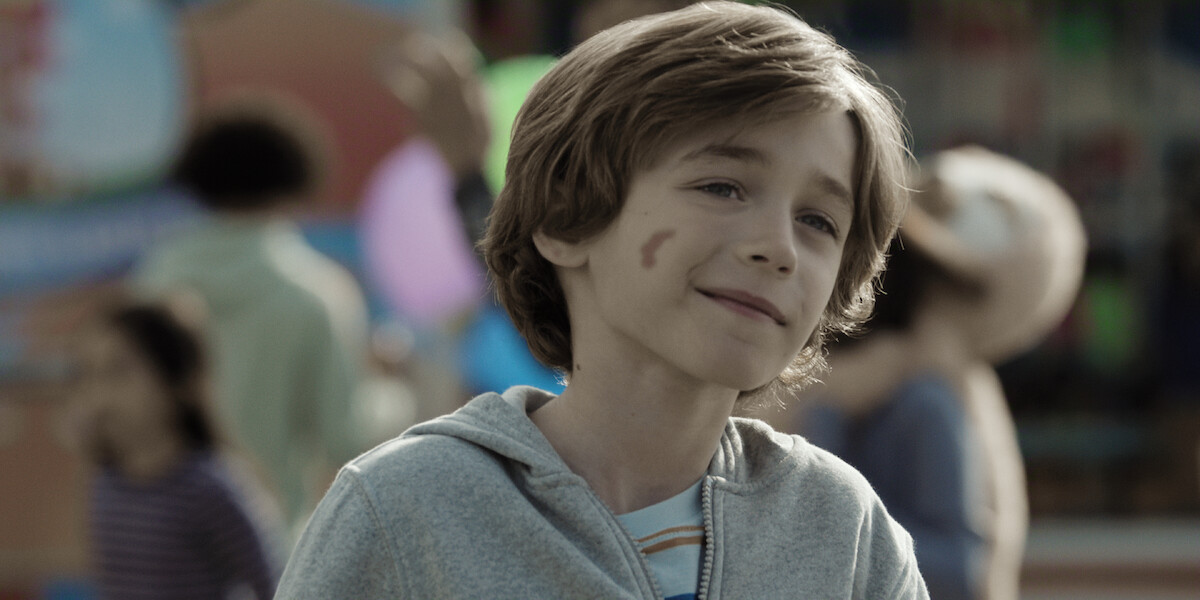 Smiling young boy with a birthmark on his cheek stands outdoors at a fair or carnival, wearing a gray hoodie, with blurred people and colorful decorations in the background.