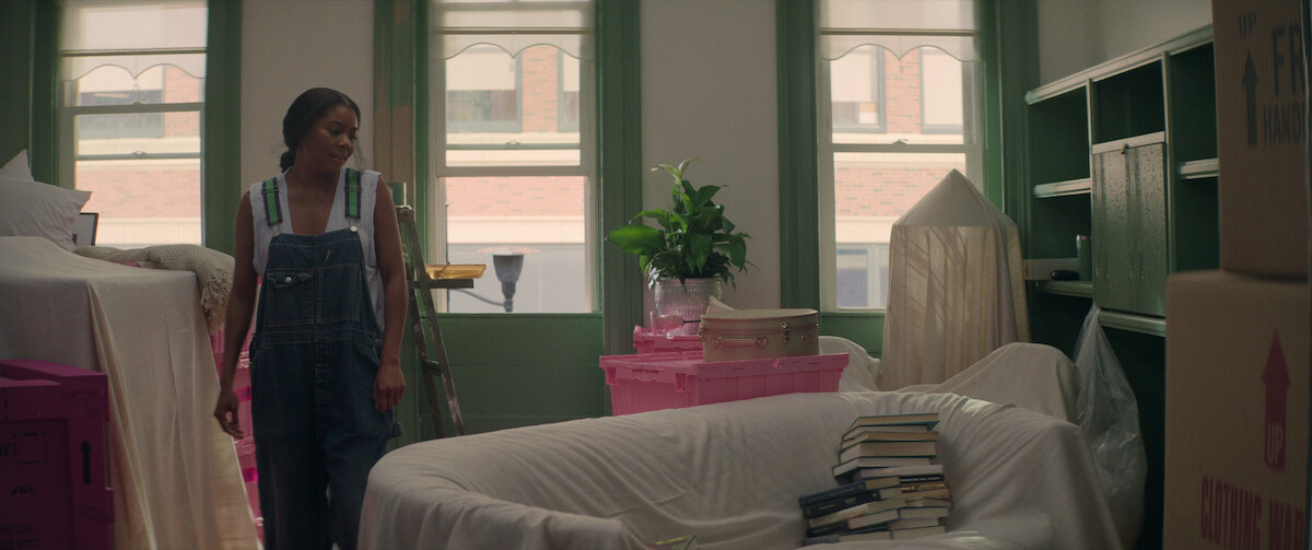 Gabrielle Union as Jenna Jones wears oversize overalls and stands next to a couch covered in a moving sheet in a still from ‘The Perfect Find.’