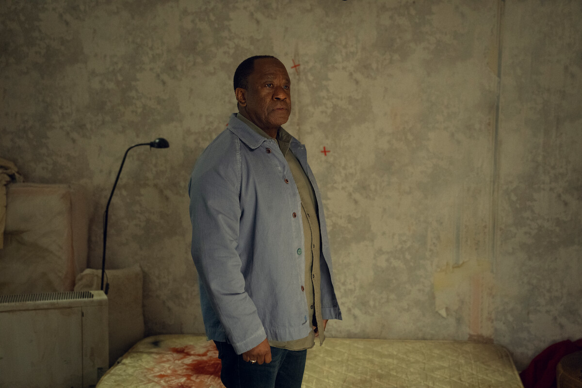A man stands in a dimly lit, run-down room with stained bedding and bare concrete walls, looking serious and contemplative. The environment appears neglected and bleak, creating a tense, somber mood.