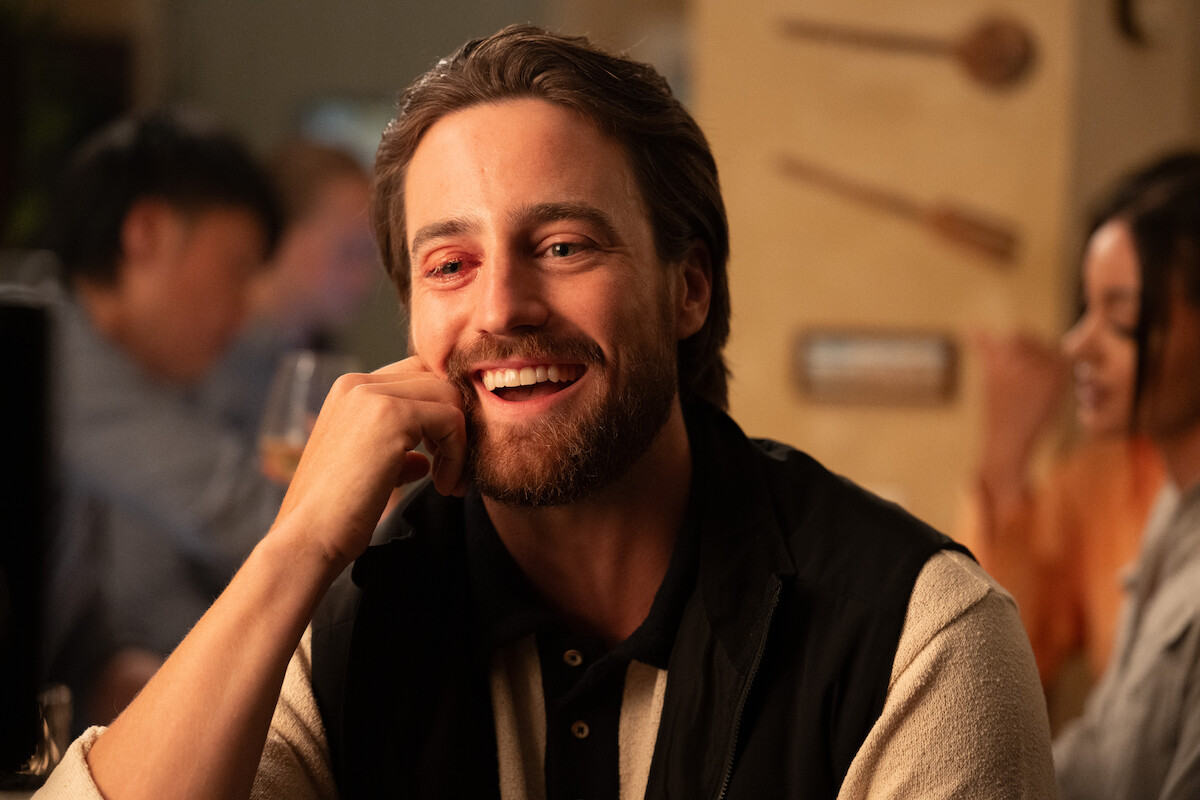 A smiling man with a reddish mark near his eye sits at a cozy, warmly lit restaurant or bar, engaging in conversation with people in the blurred background.