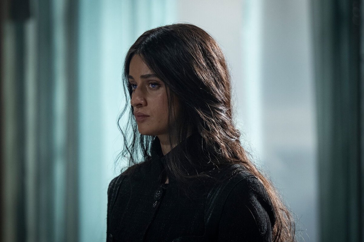 A woman with long dark hair stands indoors in soft, diffused light, wearing a dark outfit. She appears serious and contemplative, with a somber mood reflected by the muted background and calm environment.
