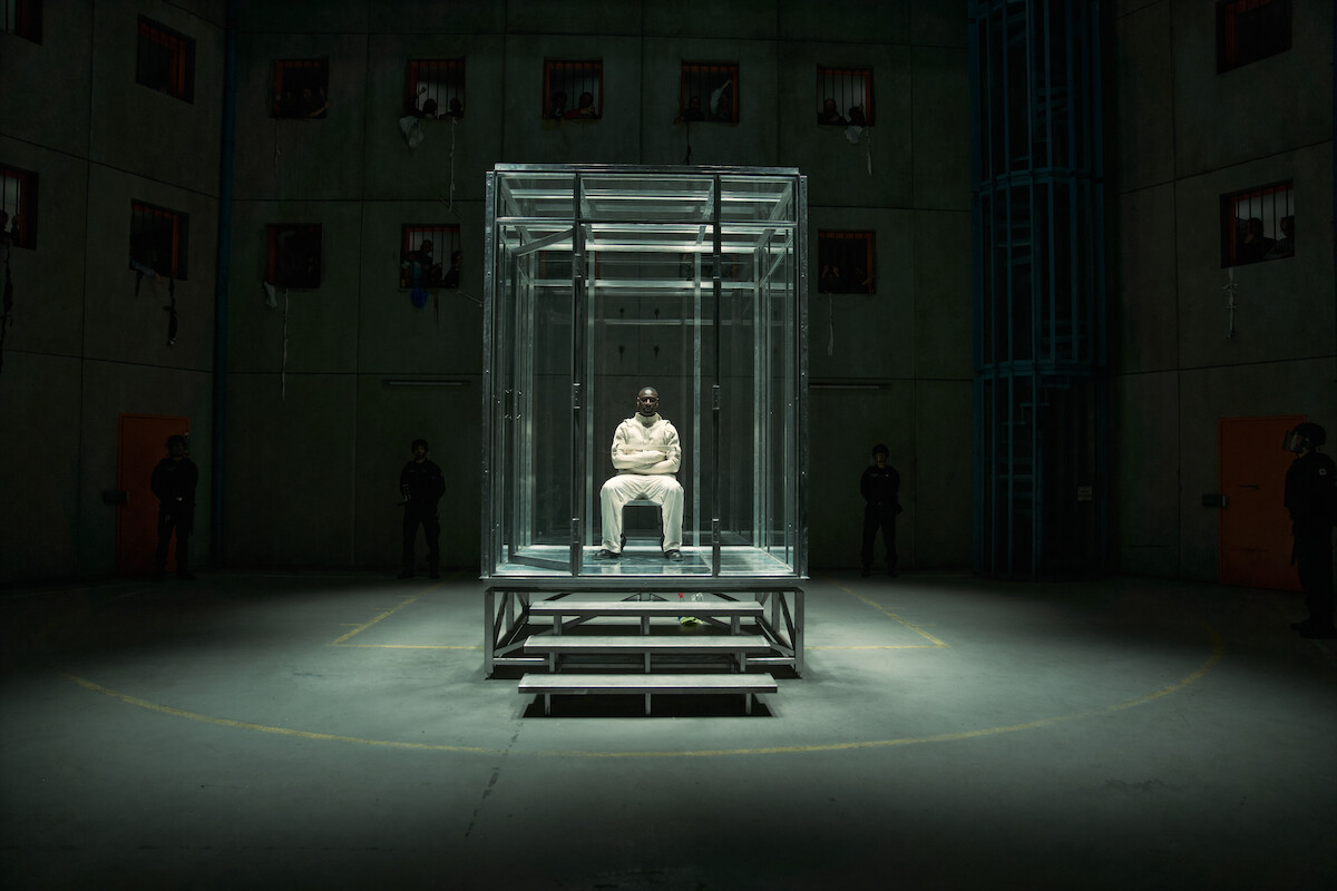 A person in a white outfit sits alone inside a glass cell at the center of a dimly lit, industrial room with concrete walls. Several shadowy figures stand in the background, emphasizing a tense, isolated prison environment.