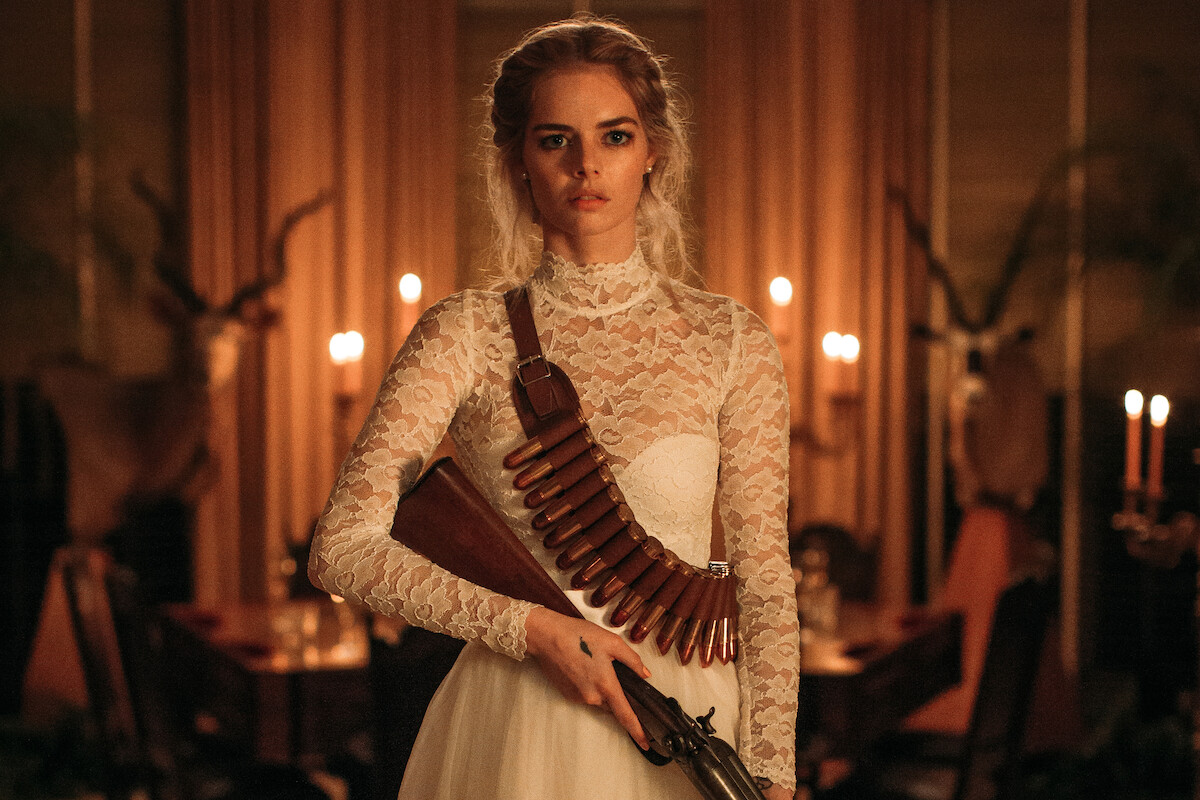 Woman in a white lace wedding dress with a shotgun and ammo belt stands in a candlelit, elegant dining room, appearing serious and determined.