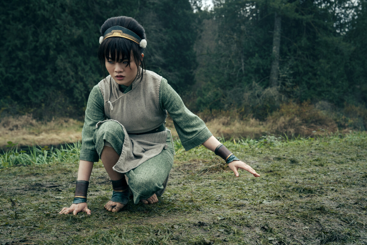 Miyako as Toph with black hair wearing green clothes crouches down in the woods with bare feet on grass and right hand touching the ground.