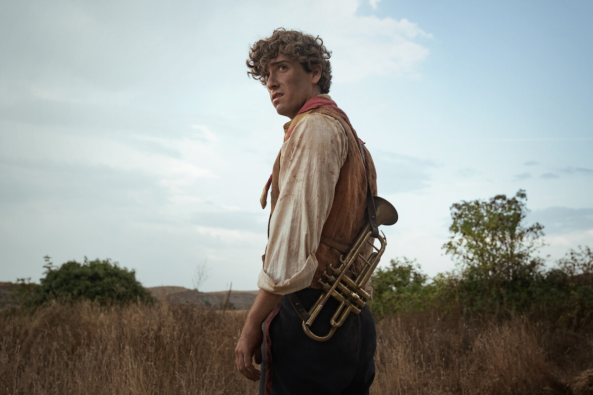 Young man in rustic, period clothing stands in a dry, grassy field at dusk, holding a brass trumpet slung over his back, with trees and distant hills under a cloudy sky in the background.
