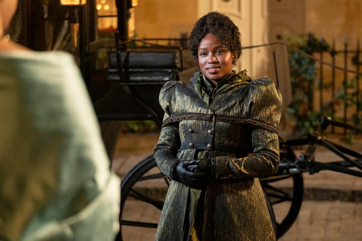 A woman in period costume stands outdoors at night in front of a horse-drawn carriage, wearing gloves and a coat, with a blurred figure and old-fashioned buildings in the background.