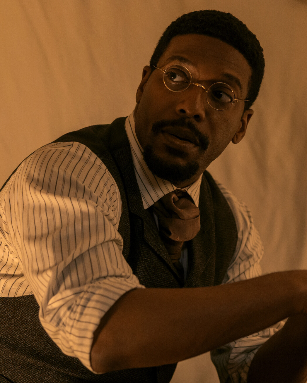 A man wearing round glasses, a striped shirt, vest, and tie sits indoors in warm, soft lighting, appearing thoughtful or in conversation, against a plain background.