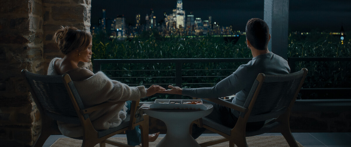 Two people sit on a balcony at night holding hands, facing a city skyline illuminated in the distance, with a small table and cozy seating, suggesting a peaceful, intimate setting.