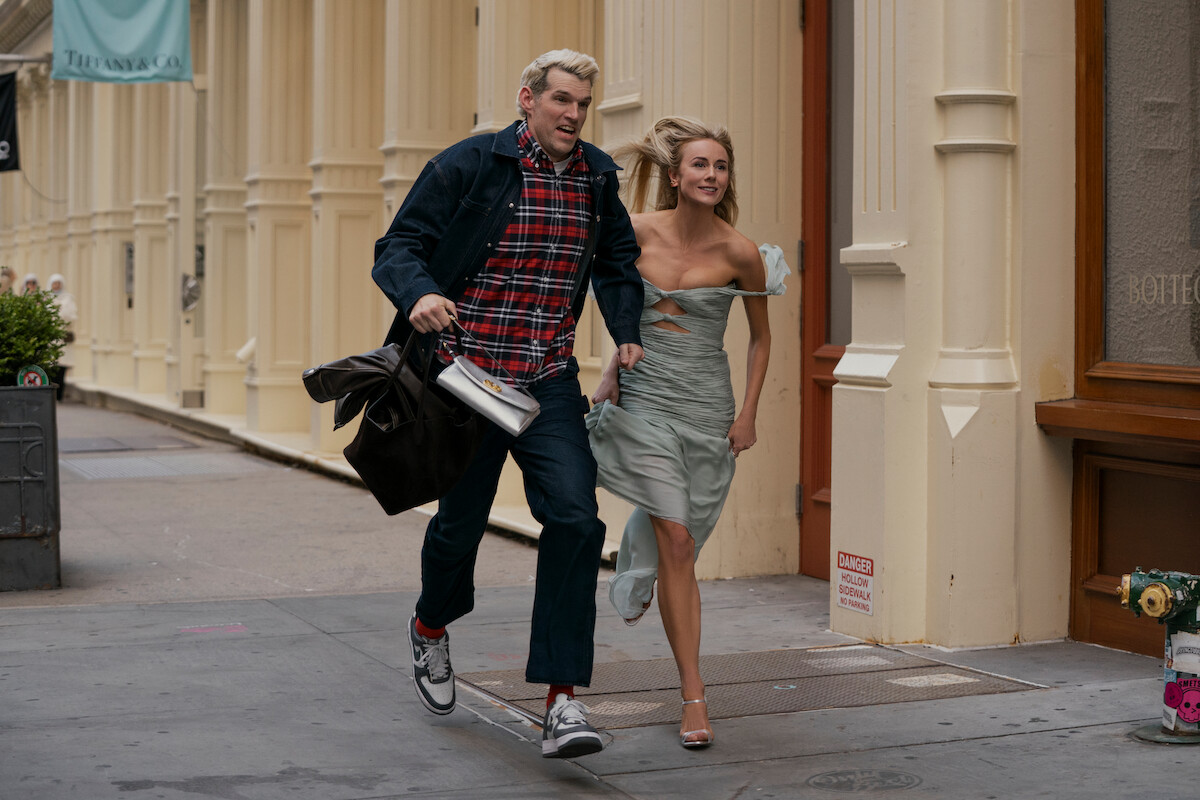 A man and woman joyfully run together on a city sidewalk, passing by cream-colored buildings with large windows. The man carries a bag and wears casual clothing, while the woman wears a light dress. Urban daytime setting.