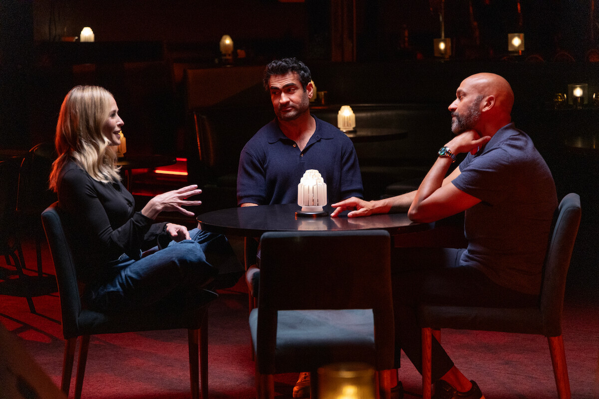Three people sit around a small round table in a dimly lit lounge or bar, engaged in conversation. The setting features dark furniture, warm lighting, and decorative candles on tables in the background.