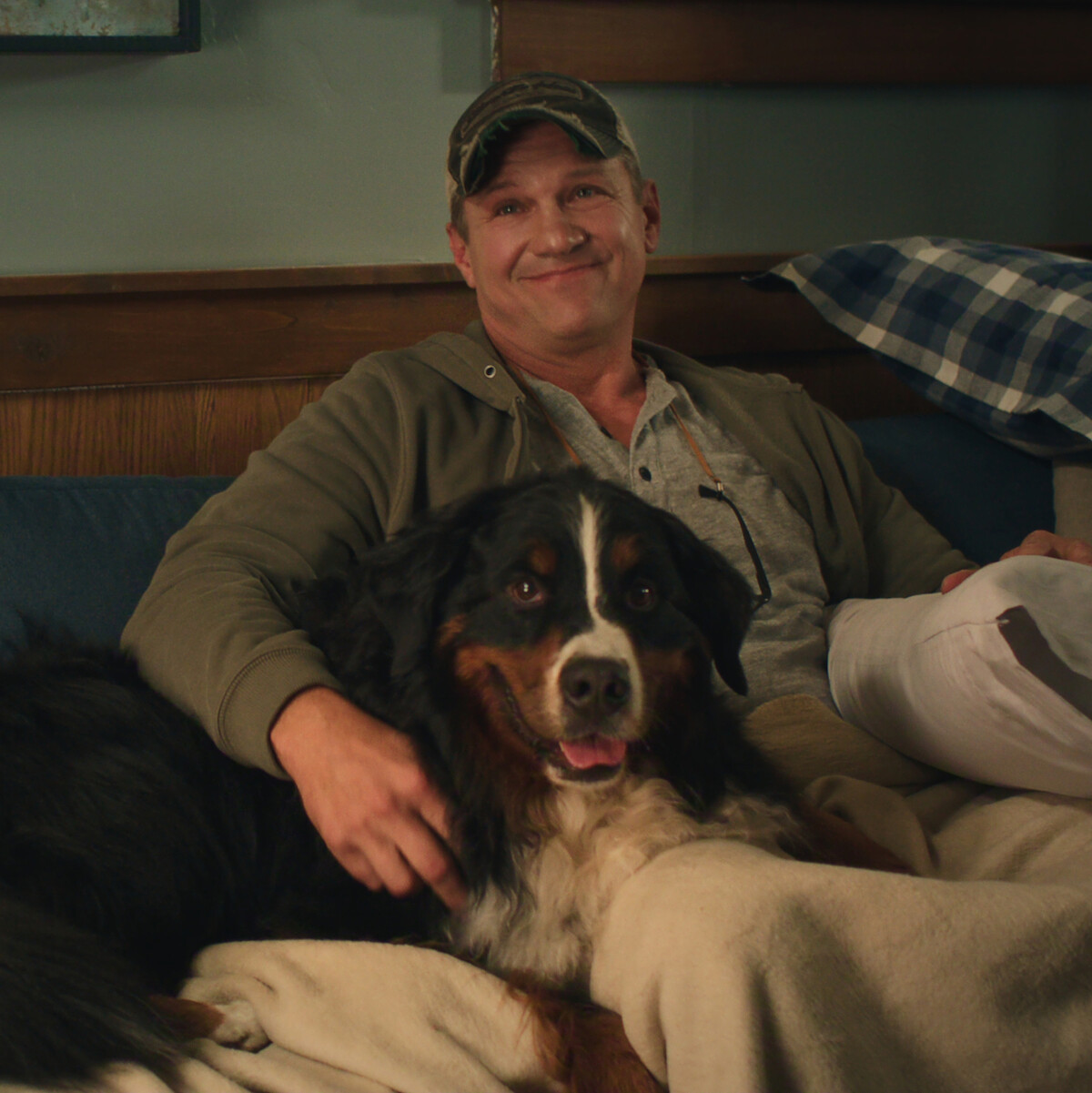 Man sitting on a couch in a cozy room, smiling while relaxing under a blanket with a large Bernese Mountain Dog, warm lighting and plaid pillows in the background.