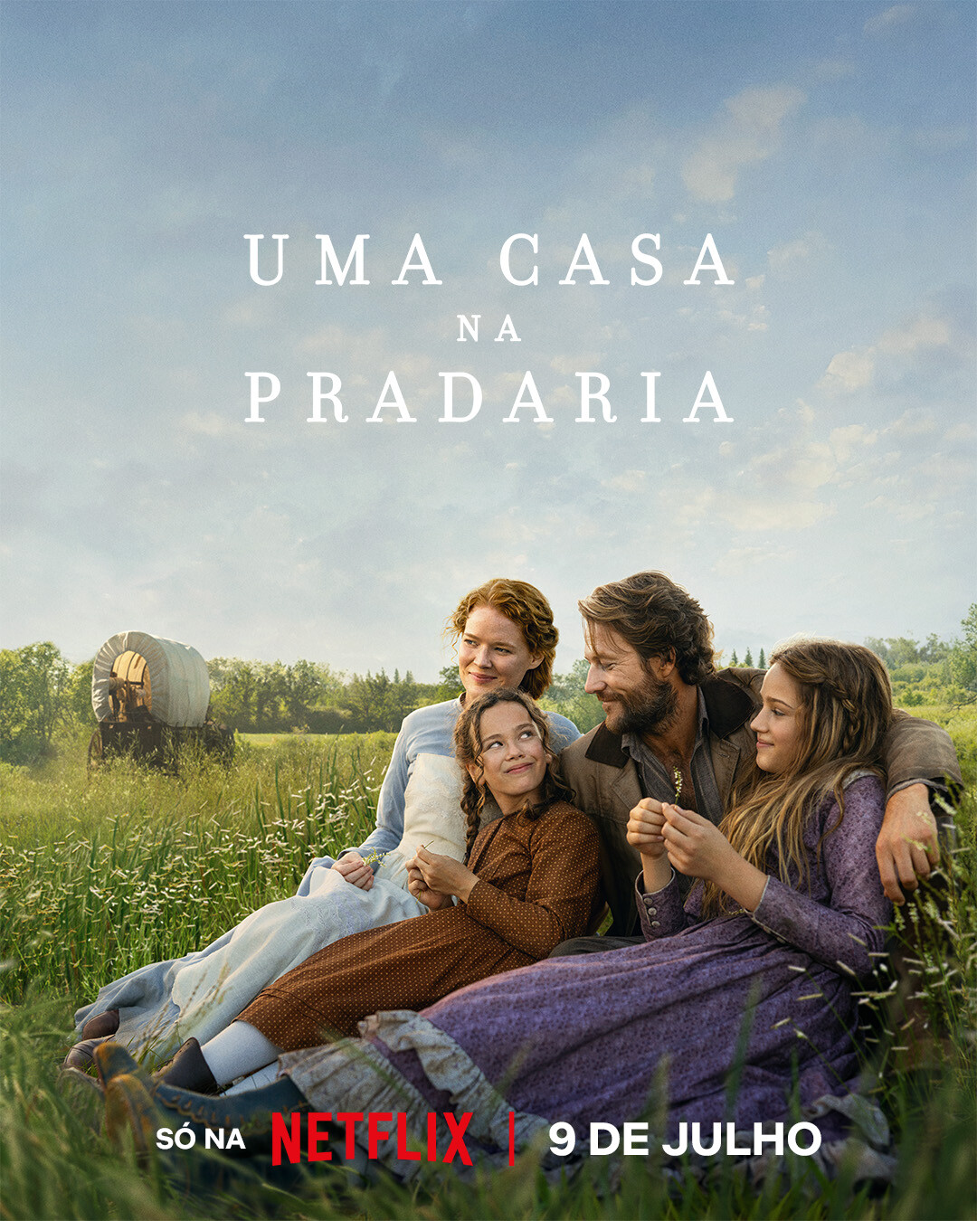 Família sorrindo ao ar livre em uma pradaria com gramado e um céu azul, ao lado de uma carroça coberta. A imagem é de ‘Uma Casa na Pradaria’.