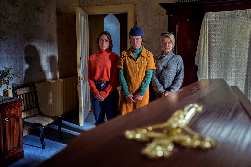 Three people stand solemnly in a dimly lit room beside a wooden casket with a gold cross, featuring vintage wallpaper, a chair, and a wardrobe, suggestive of a funeral or wake in a traditional home setting.