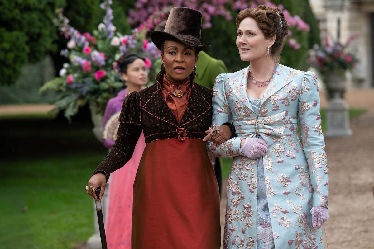 Two women in elegant period costumes walk arm in arm outdoors, surrounded by lush gardens and large floral arrangements, with other people in the background, in a historical setting.