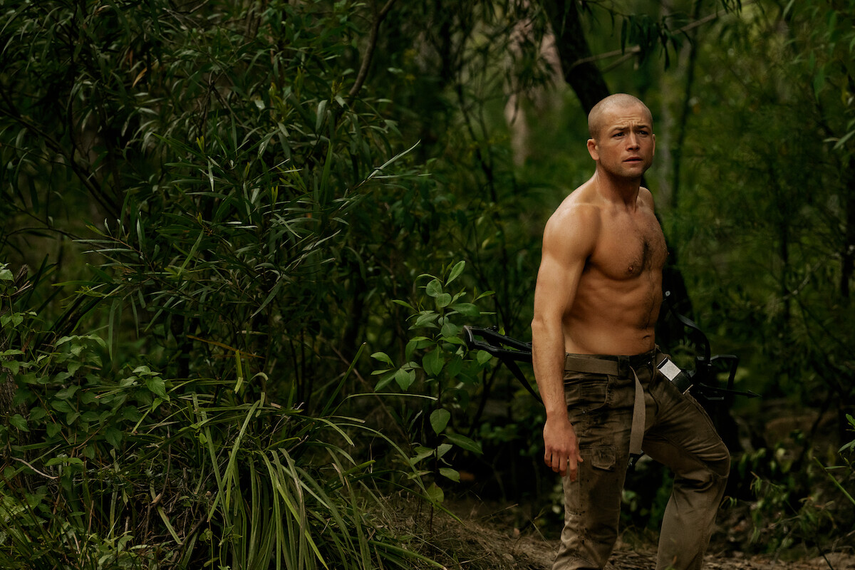 Shirtless man with short hair and muscular build holding a compound bow stands in a dense green forest, suggesting a survival or hunting scenario in natural, rugged outdoor surroundings.