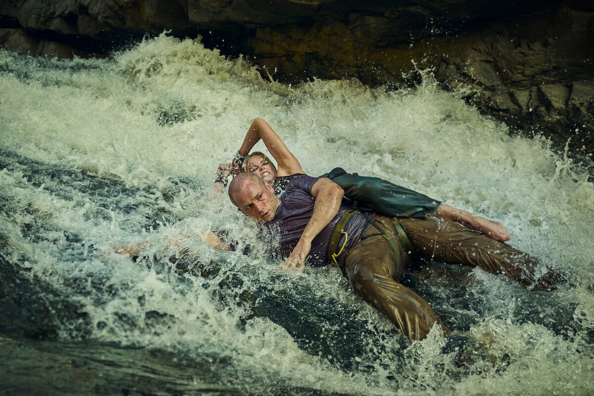 Two people in wet clothing lie intertwined on a rocky riverbed with fast-moving water, set against natural rocks. The scene feels urgent and dramatic, evoking a sense of struggle or survival in a wild environment.Edit Alt Text