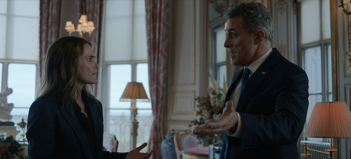 Two people in formal attire are having an intense conversation in an elegant, well-lit room with large windows, ornate decor, drapes, and lamps, suggesting a serious discussion in a high-end or official setting.