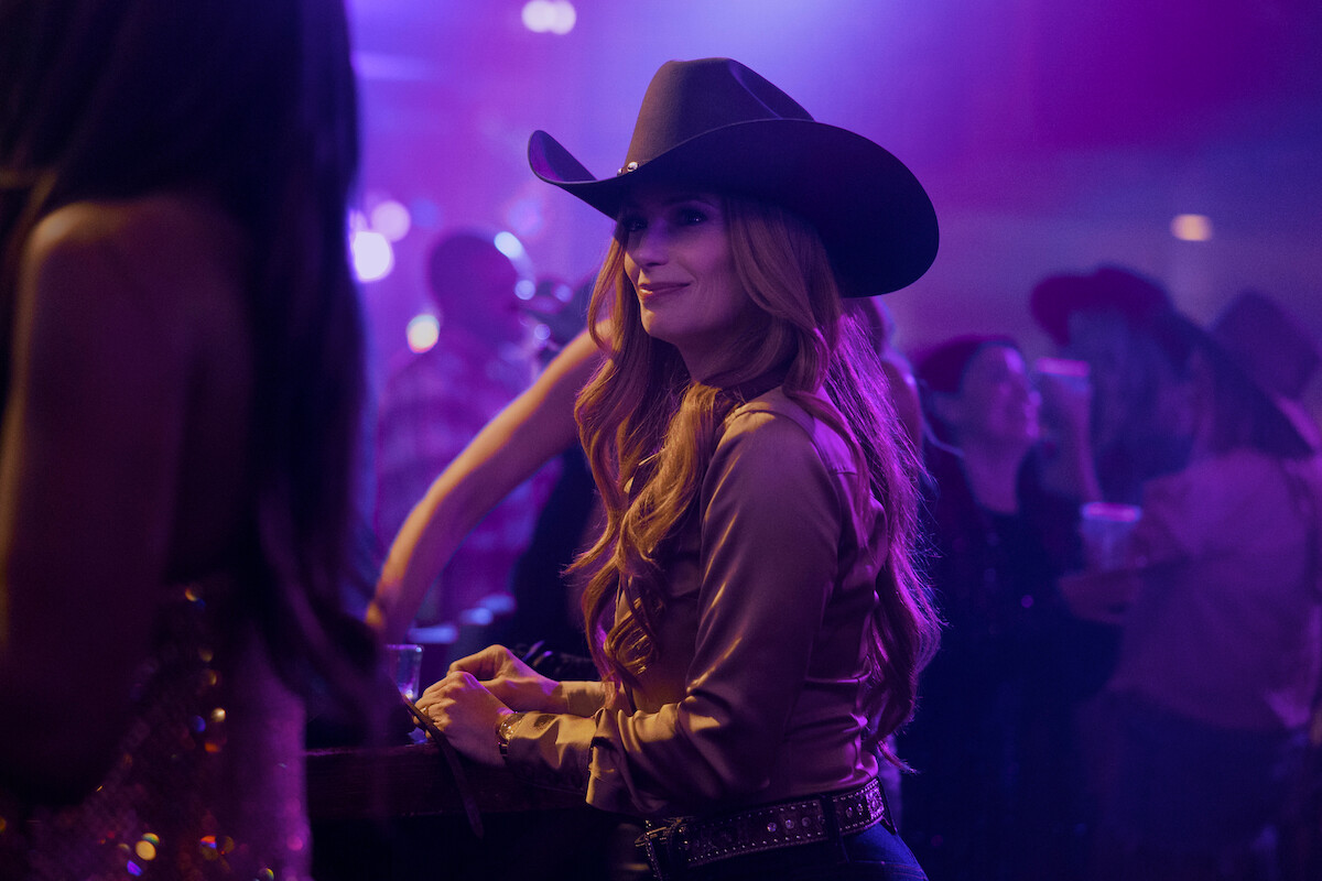 Mulher usando um chapéu de caubói e uma camisa reluzente em uma balada ou bar country animado com pouca iluminação, pessoas em segundo plano, luzes coloridas e um clima de festa.
