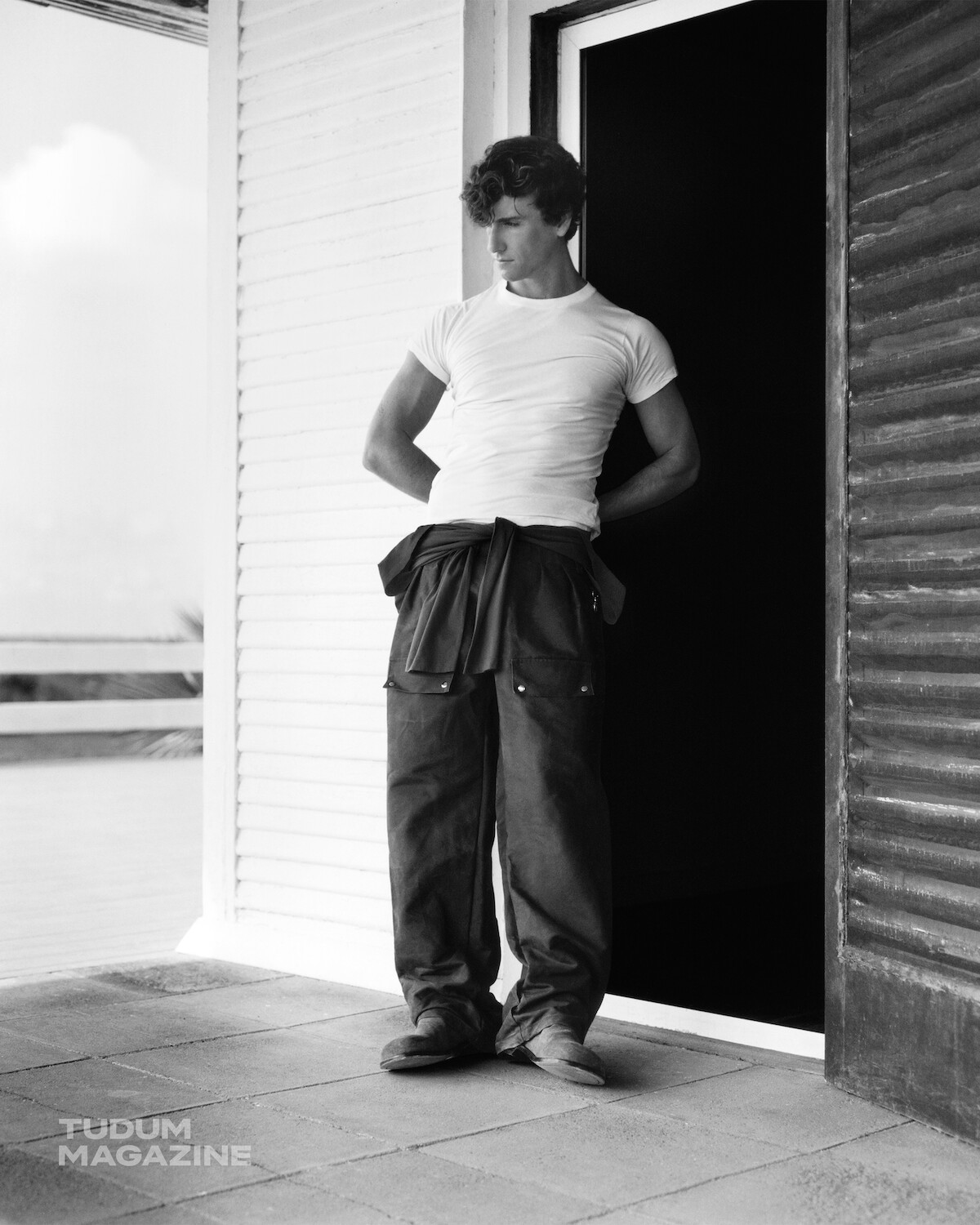Man in white t-shirt and work pants stands in front of an open door on a porch, looking down with hands behind back. The setting appears to be outdoors with a modern building and railing visible in the background. Black and white photo.