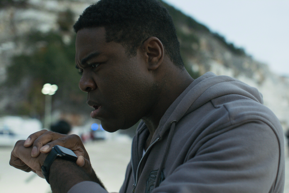 Man in a gray hoodie outdoors intensely looks at smartwatch, with blurred background featuring cliffs, trees, and distant vehicles with lights, suggesting an urgent or focused situation.