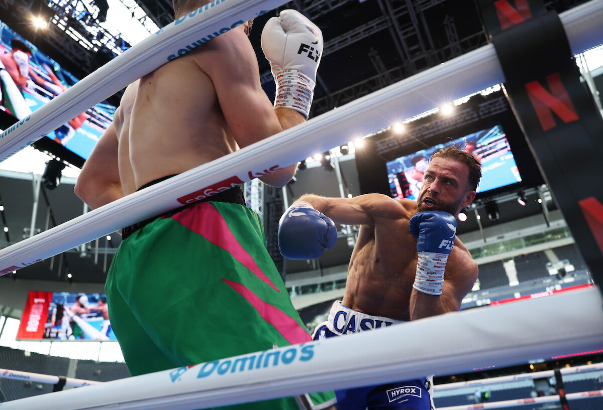 Two boxers fighting in a brightly lit boxing ring inside a modern stadium, seen from a low angle outside the ropes, with screens and advertisements visible in the background.