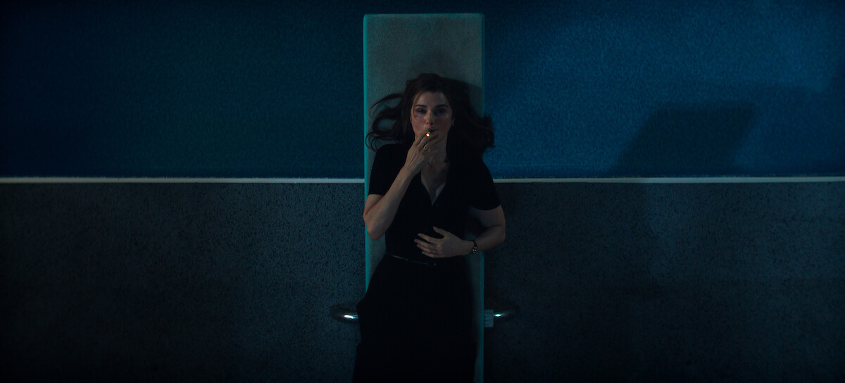 Rachel Weisz as M lying on a diving board in a dimly lit indoor pool area, wearing dark clothes, with her hand covering her mouth, and blue walls in the background.