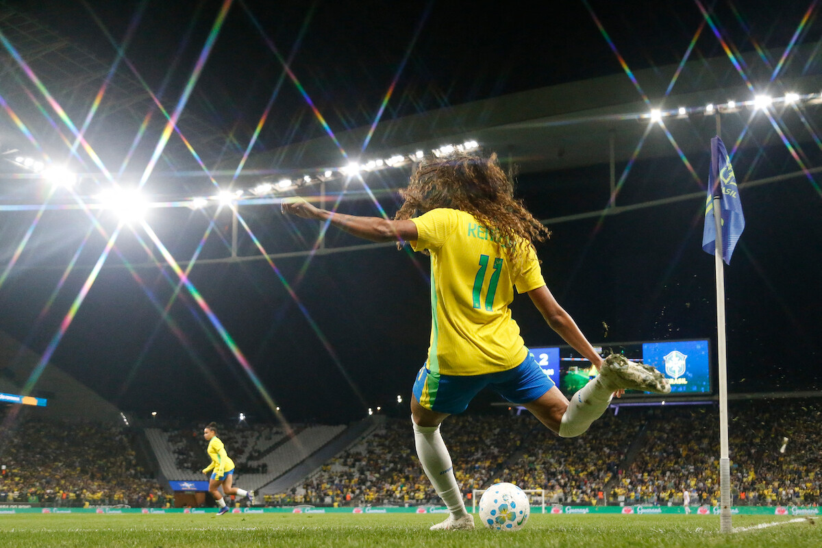 Soccer player in yellow jersey taking a corner kick at night in a stadium, with bright lights, a crowd of spectators, and other players visible in the background.