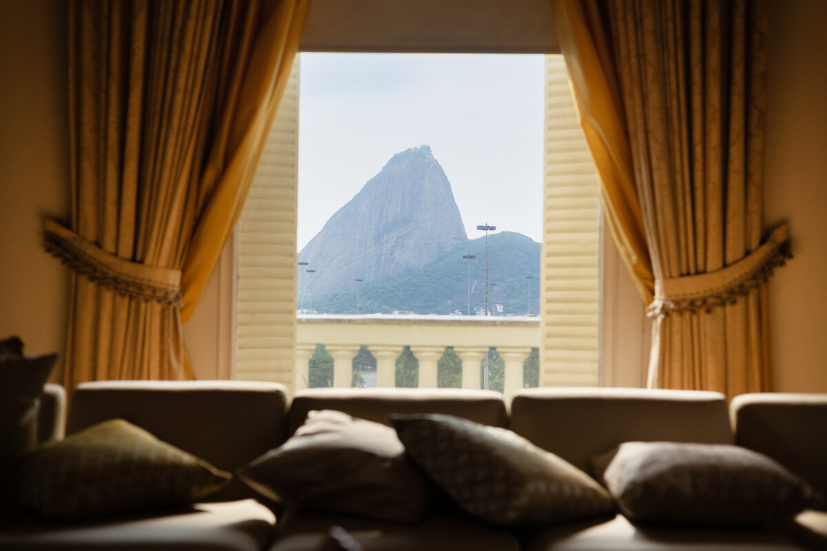 Olhando de dentro de uma sala é possível ver um sofá com almofadas, em primeiro plano. Atrás dele tem uma cortina amarela e uma janela aberta. Na janela, é possível ver o Pão de Açúcar, no Rio de Janeiro