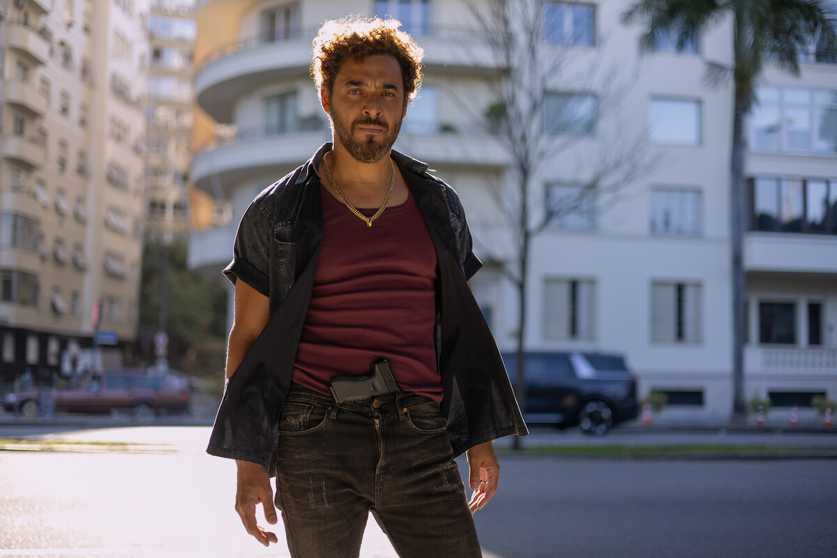 Um homem de meia idade tem cabelos e barba curtos e veste camisa azul sobre camiseta vinho e calça. Ele leva uma arma de fogo presa ao cinto