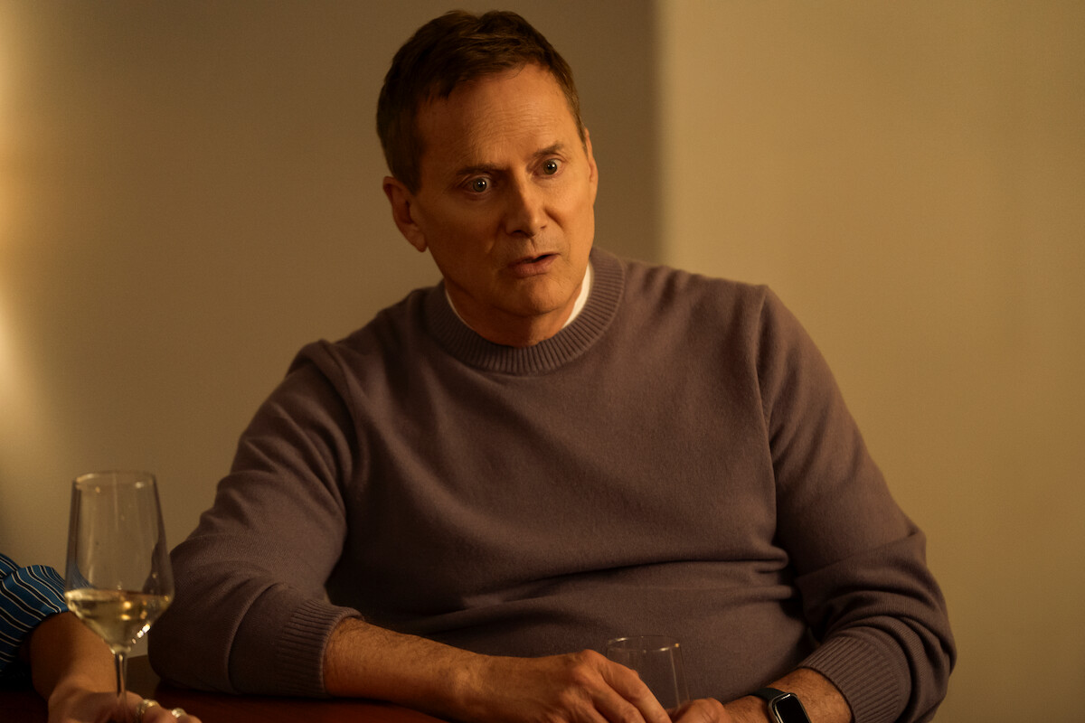Man in a purple sweater sitting at a table in a warmly lit indoor setting, looking engaged in conversation, with a wine glass and another person partially visible nearby.