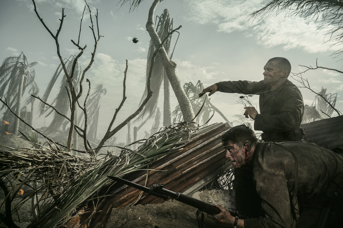 Two soldiers in muddy uniforms take cover behind debris in a war-torn, smoky battlefield with broken trees, holding rifles and looking alert, surrounded by chaos and destruction.