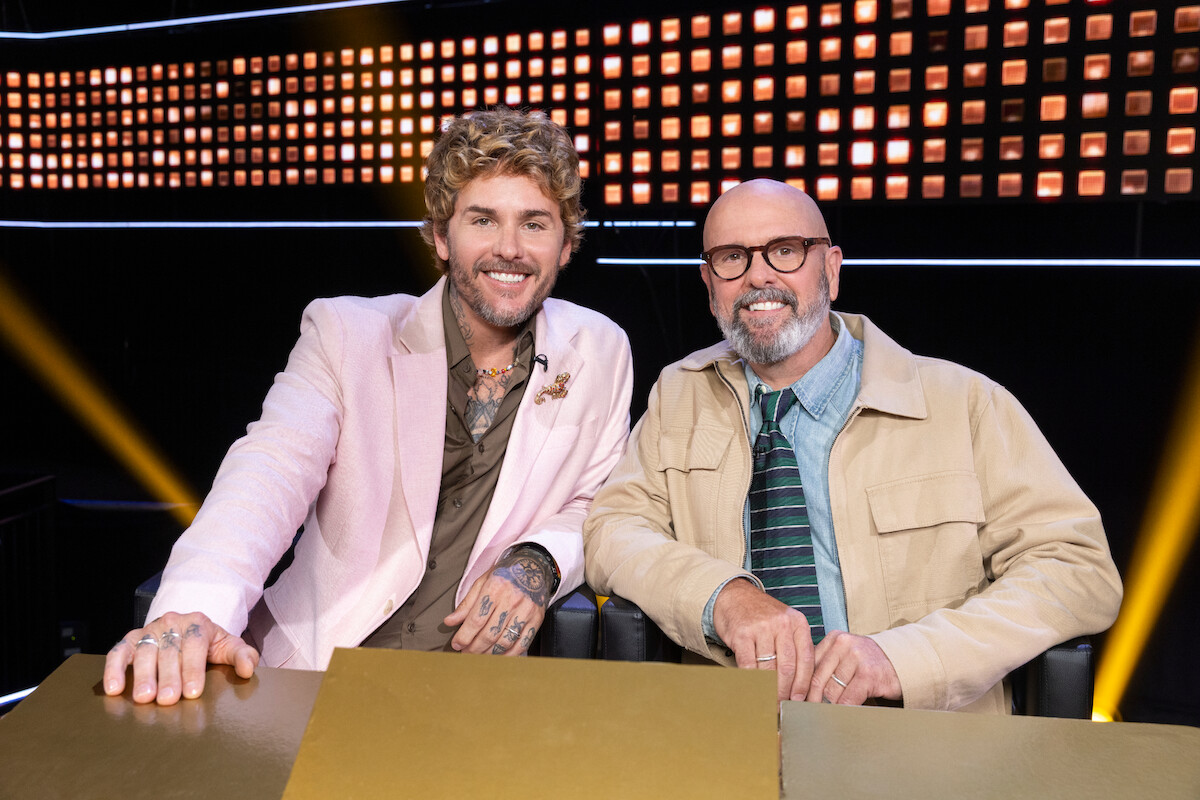 Two smiling men sit together behind a gold desk in a brightly lit studio with a modern, illuminated backdrop, suggesting a lively and welcoming atmosphere, possibly on a TV show set or competition panel.
