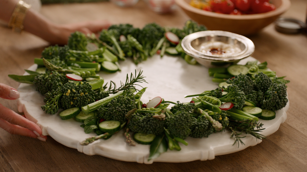 Vegetable wreath with broccoli, cucumber, radish, and herbs arranged on a marble platter with a dip in a silver bowl, set on a wooden table in a cozy, inviting kitchen environment.
