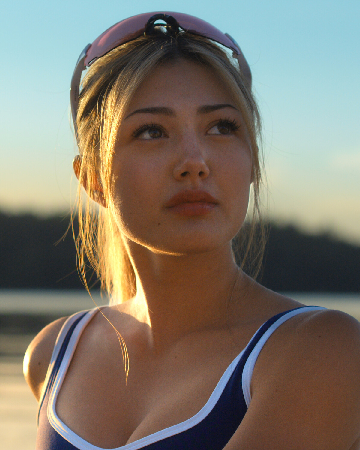 Woman in athletic wear looking into distance outdoors at sunset, with sunglasses on her head, soft light on her face, and a blurred lake and treeline in the background.