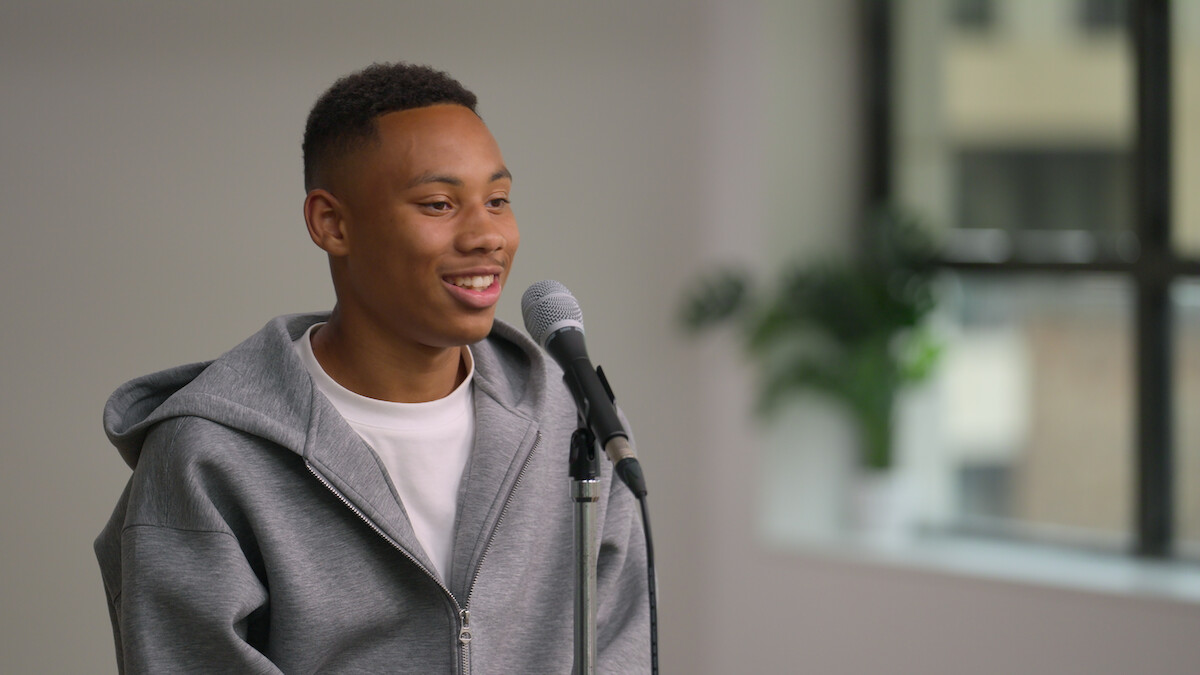 Young person in a gray hoodie smiles while speaking into a microphone in a bright, modern indoor setting with a large window and green plant in the background, creating a relaxed and positive mood.