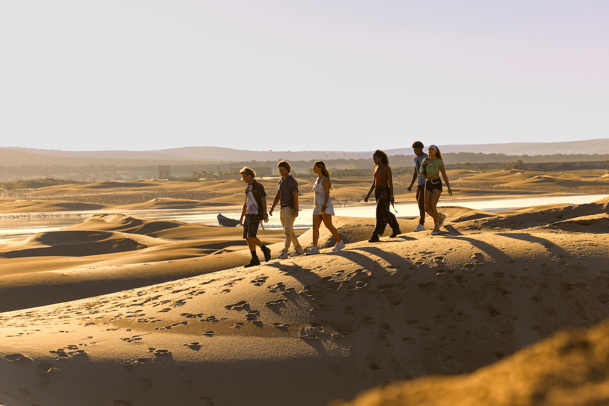 Group of six people walking on sandy desert dunes during daylight, with long shadows and distant water, under a clear sky, surrounded by a wide, open landscape.