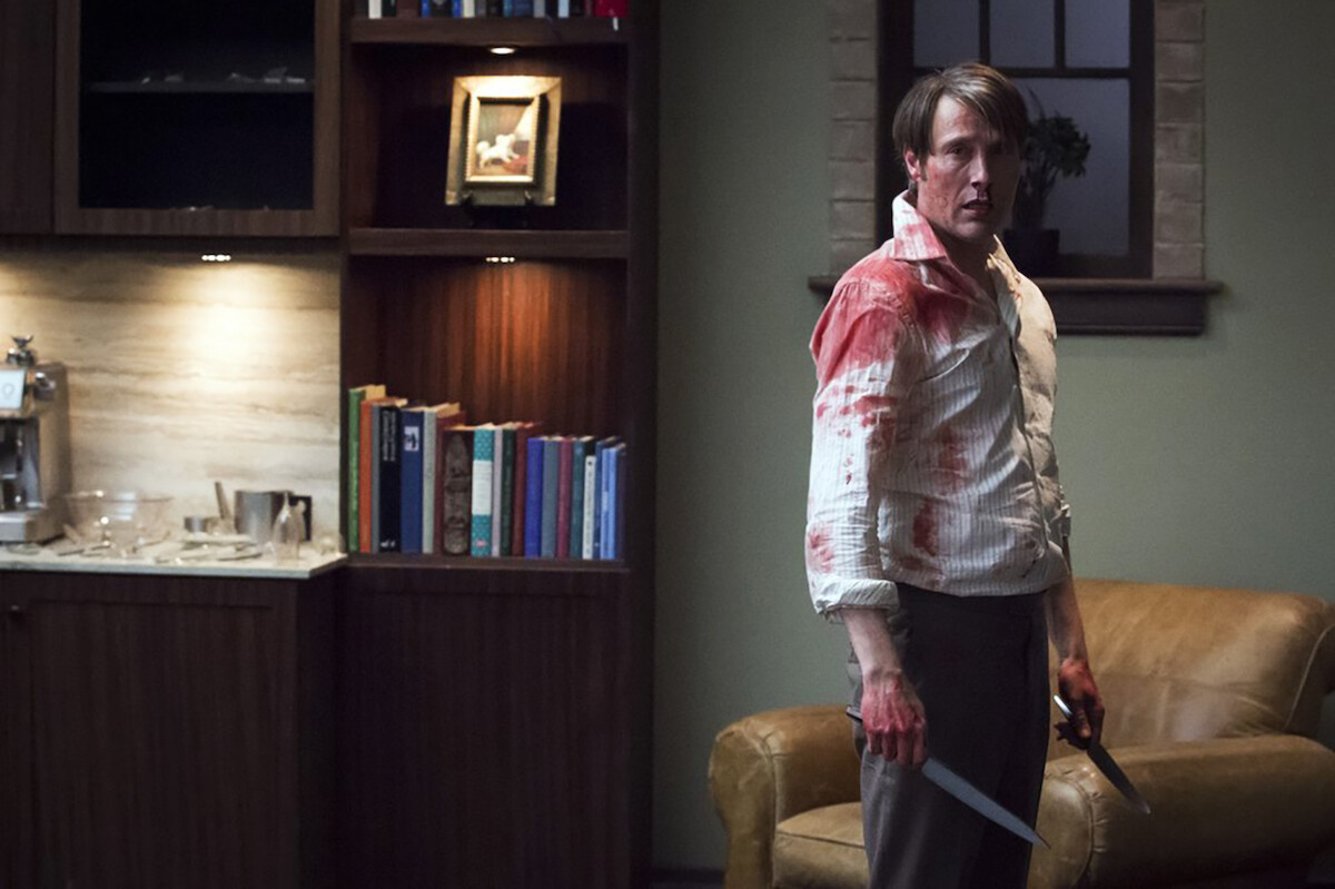 A man with bloodstained clothes holds two knives, standing in a dimly lit living room with a leather couch, bookshelves, window, and kitchen counter in the background.