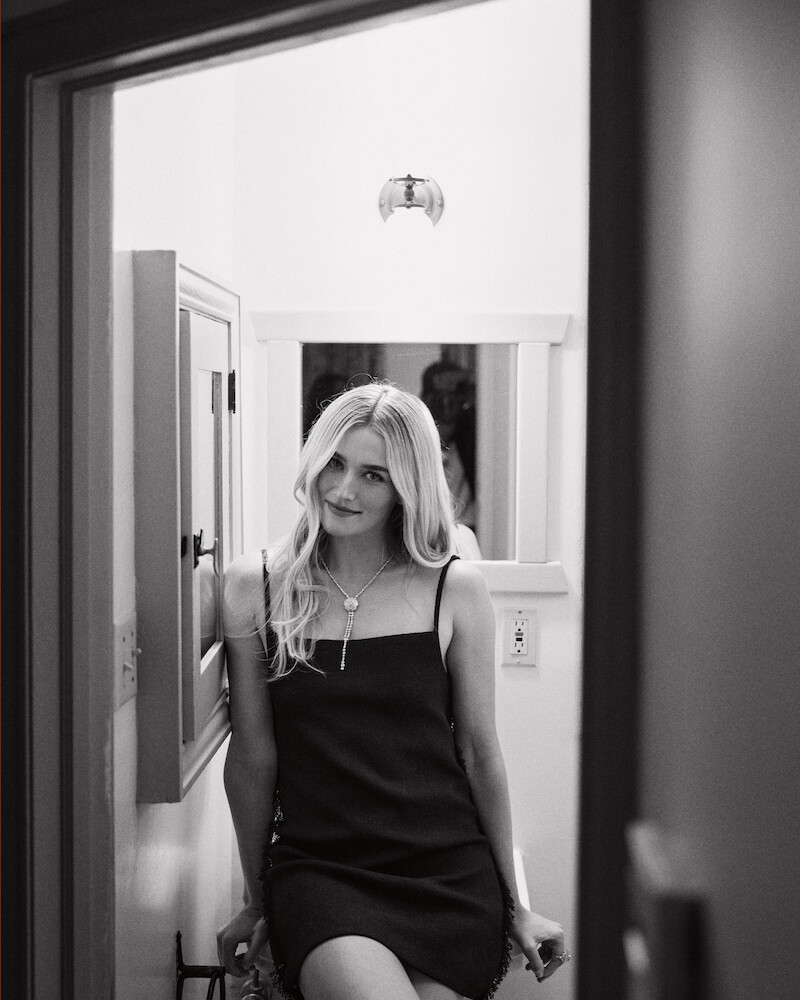 A woman with long blond hair in a black dress sits on a counter in a small, bright bathroom. The scene is framed by a door, with a mirror and wall fixtures visible in the background. Black-and-white photograph.